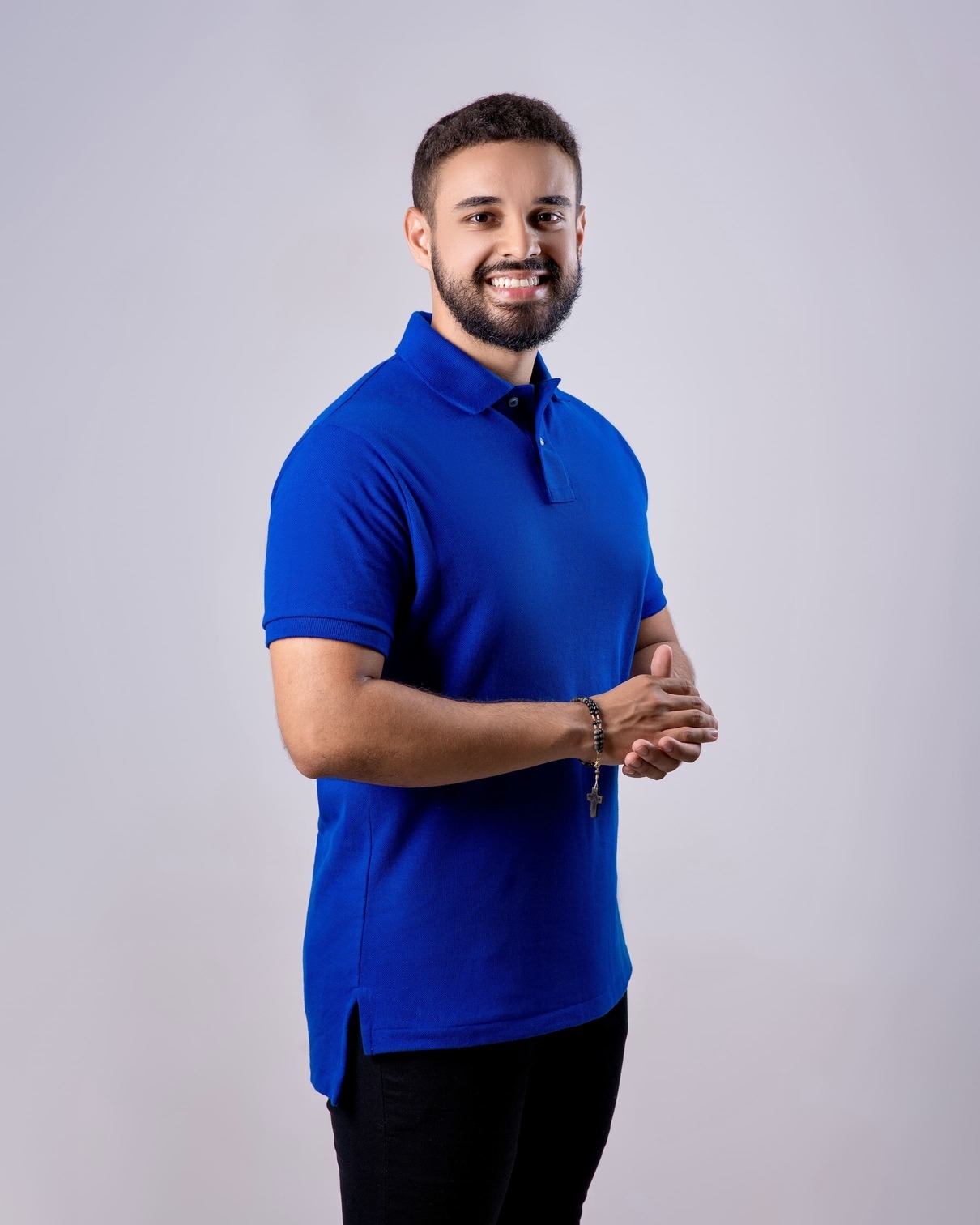 Foto de Luciano Ribeiro, homem pardo, com barba e olhos e cabelo castanhos, vestindo uma camisa do tipo polo de cor azul, com uma mão sobre a outra próximas a barriga, olhando fixadamente para a câmera e sorrindo.